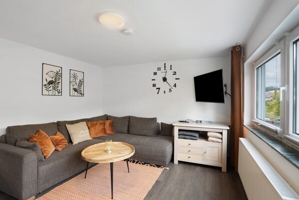 A modern living room with a grey sofa, a round wooden coffee table, a white cabinet, a mounted TV, and a large window.