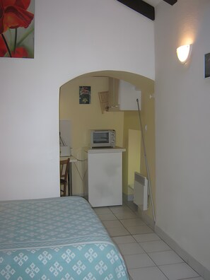 Um quarto pequeno com uma cama, um espaço de cozinha, micro-ondas, uma cadeira e um quadro na parede.