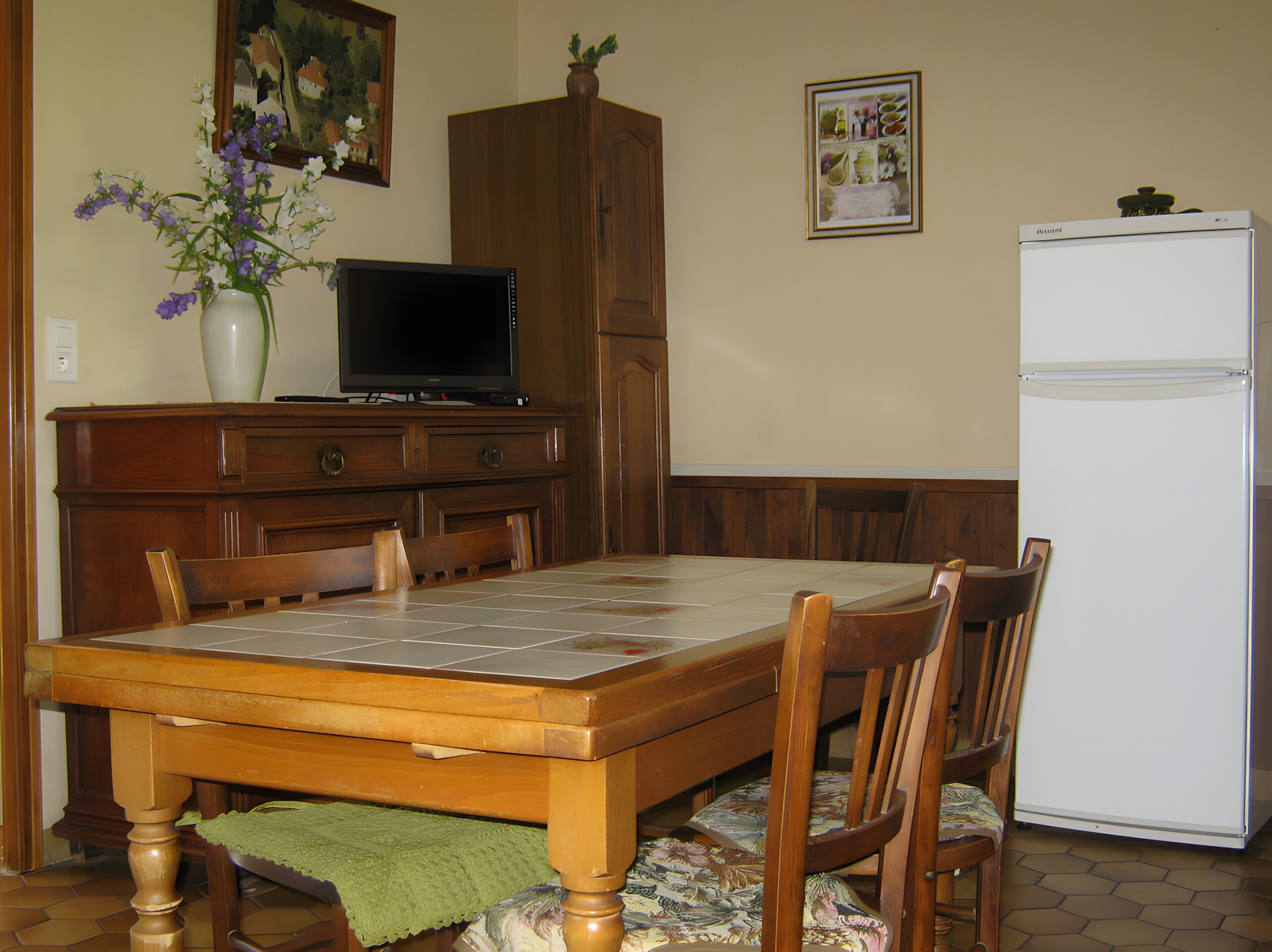 A dining area with a wooden table, chairs, a television, a refrigerator, and a vase with flowers.
