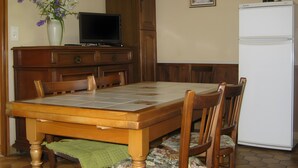 A dining area with a wooden table, chairs, a television, a refrigerator, and a vase with flowers.