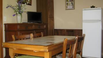 A dining area with a wooden table, chairs, a television, a refrigerator, and a vase with flowers.