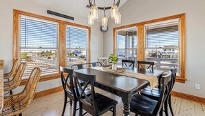 A dining area with a wooden table and chairs, large windows with blinds, and a chandelier. - Spacious KDH Getaway with Pool, Near Beach (Kill Devil Hills)