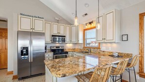 A modern kitchen with a central island, bar stools, stainless steel appliances, and a 'Welcome to the Beach' sign. - Spacious KDH Getaway with Pool, Near Beach (Kill Devil Hills)