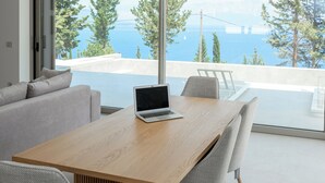 A modern living room with a wooden table, a laptop, and a sofa. A large window offers a view of the sea and trees.