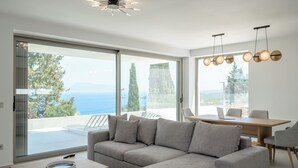 A modern living room with a grey sectional sofa, a wooden coffee table, and a dining area with a table and chairs. Large windows offer a view of the sea and trees.