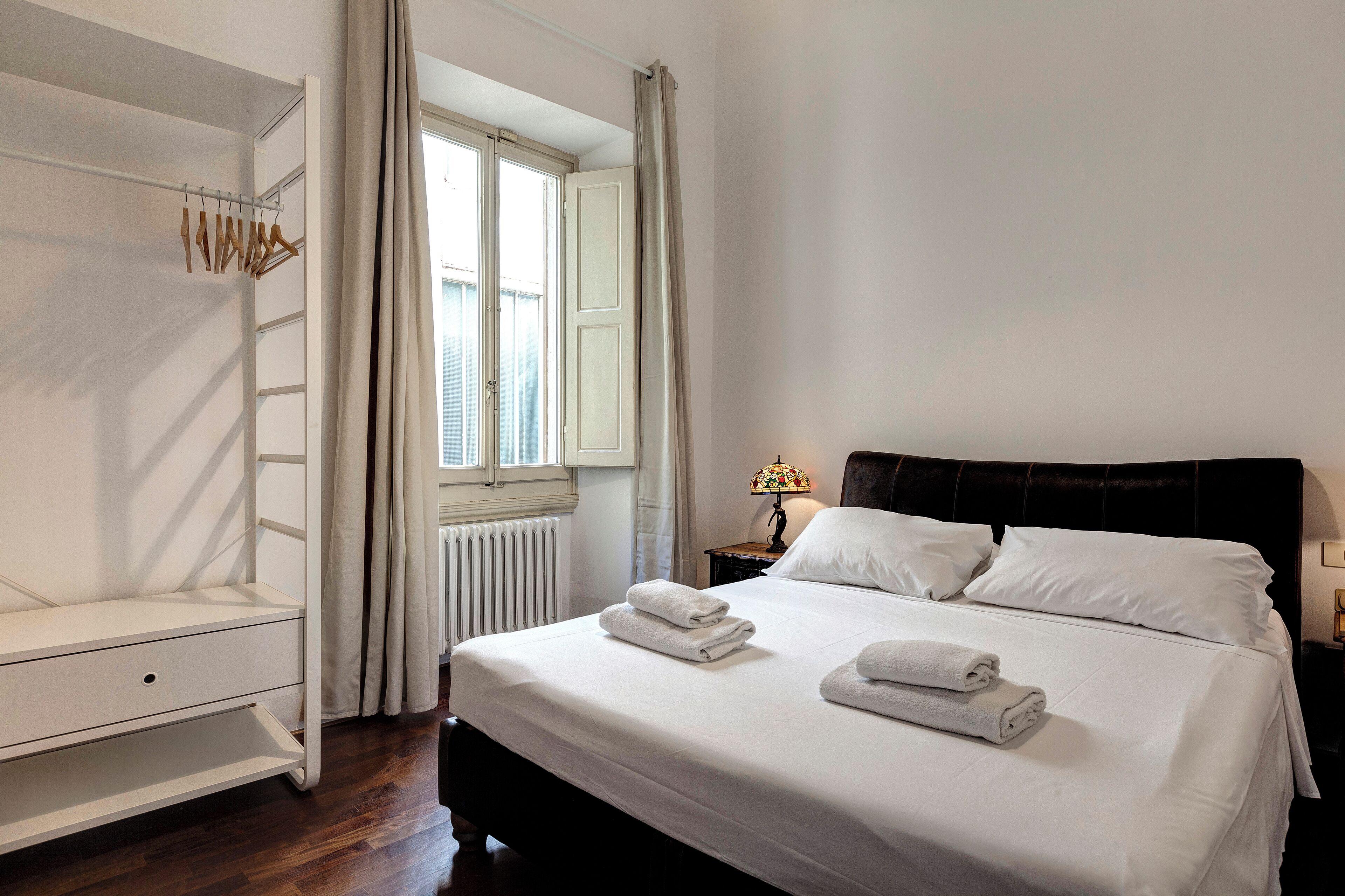 A neatly made bed with white linens and towels, a white wardrobe with a hanging rod, a window with curtains, and a bedside table with a lamp.