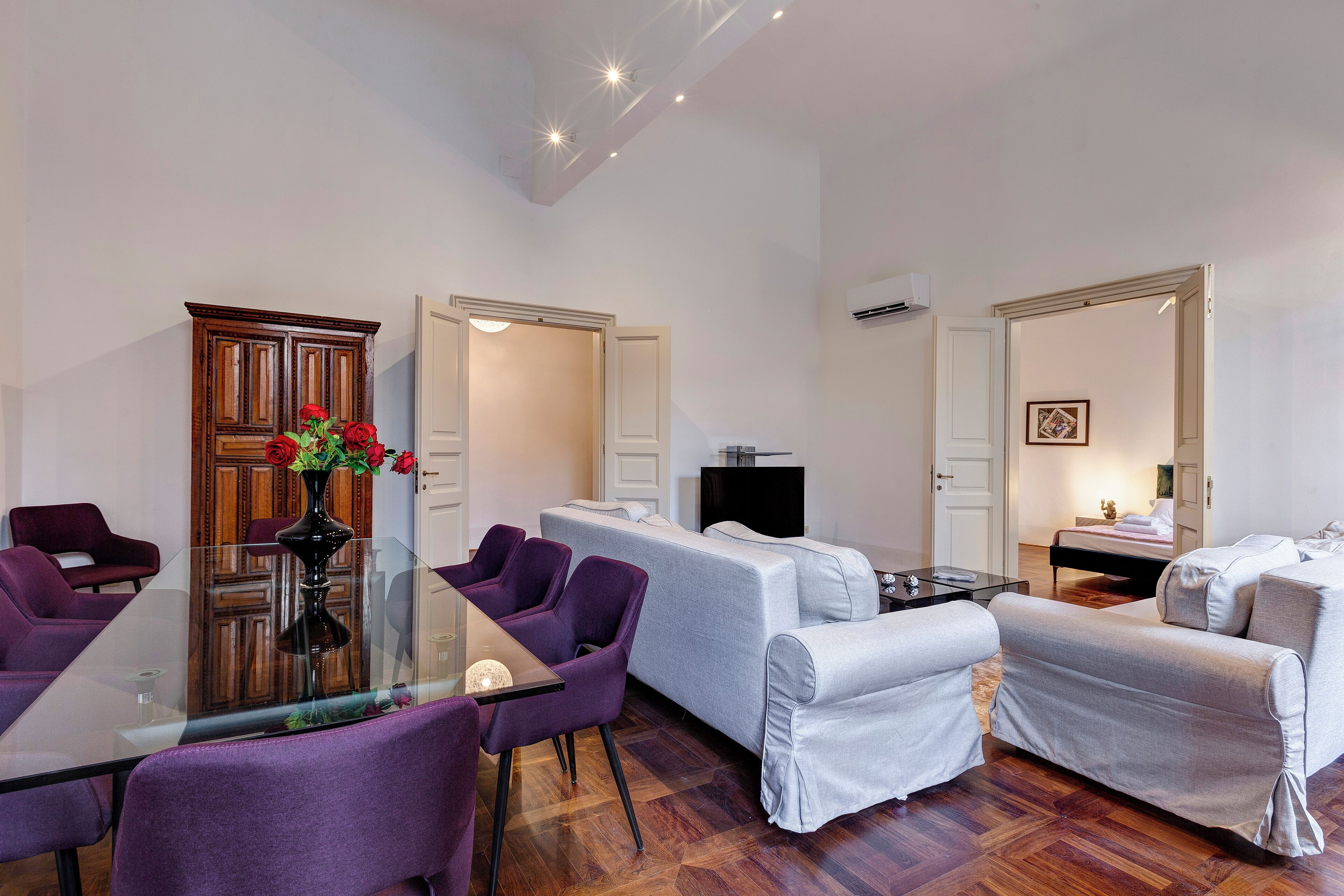 A modern living room with a glass dining table, purple chairs, a grey sofa, and a white covered sofa.