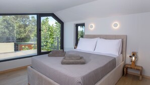 A modern bedroom with a large bed, a wooden nightstand, and a view of greenery through a large window.