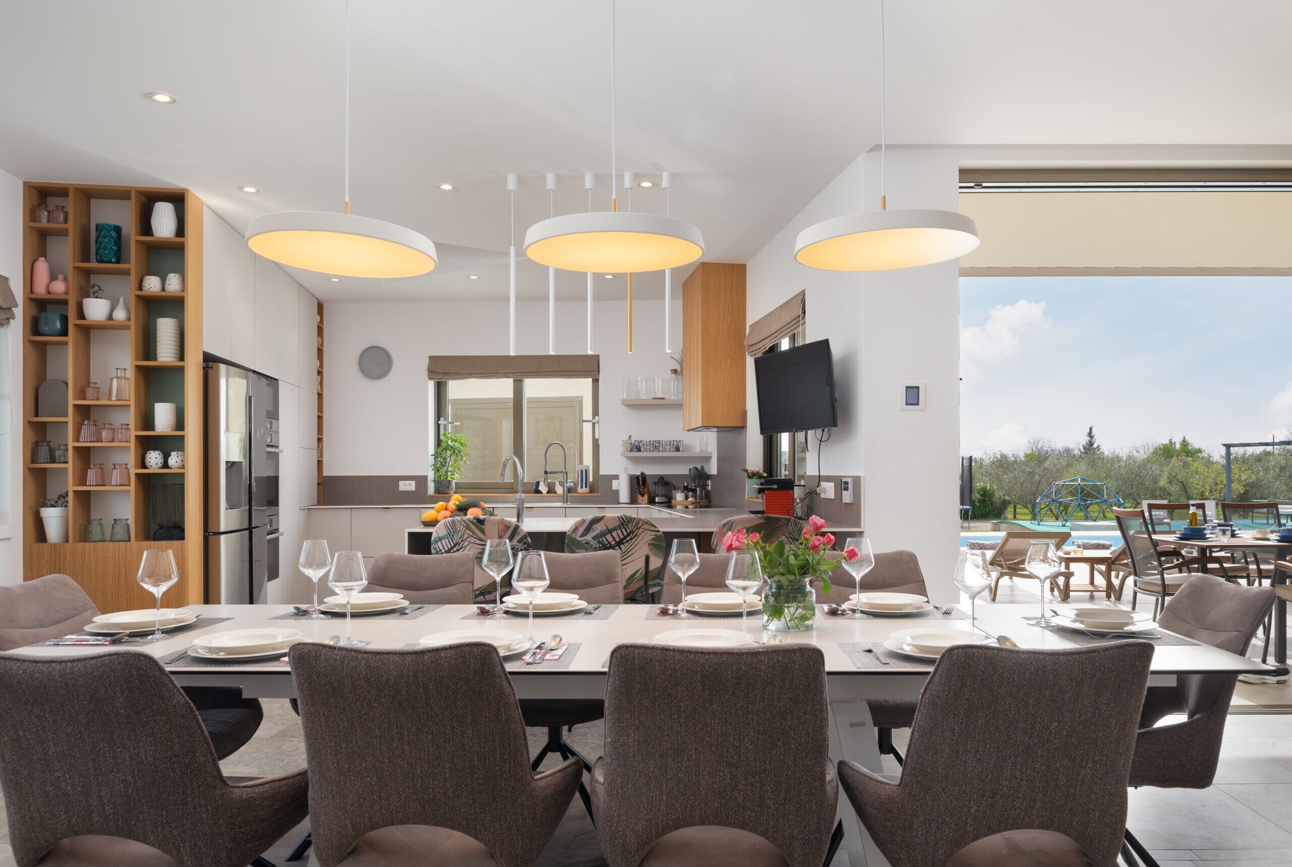 Un espace de repas moderne, avec une grande table dressée pour six personnes, entourée de chaises confortables. On aperçoit à l’arrière-plan une cuisine dotée d’une grande fenêtre et d’un téléviseur à écran plat.