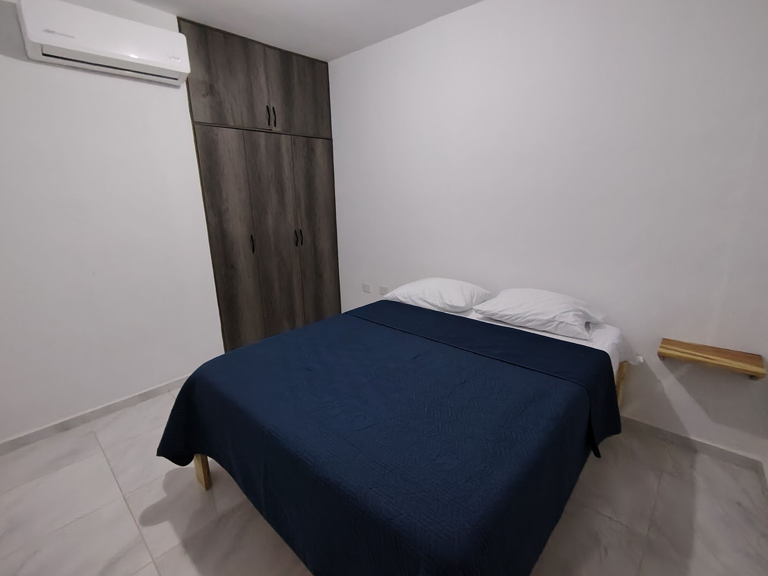 Ein Schlafzimmer mit einem Bett, weißen Kissen, einer dunkelblauen Bettdecke, einem Holzschrank und einer Klimaanlage an der Wand.