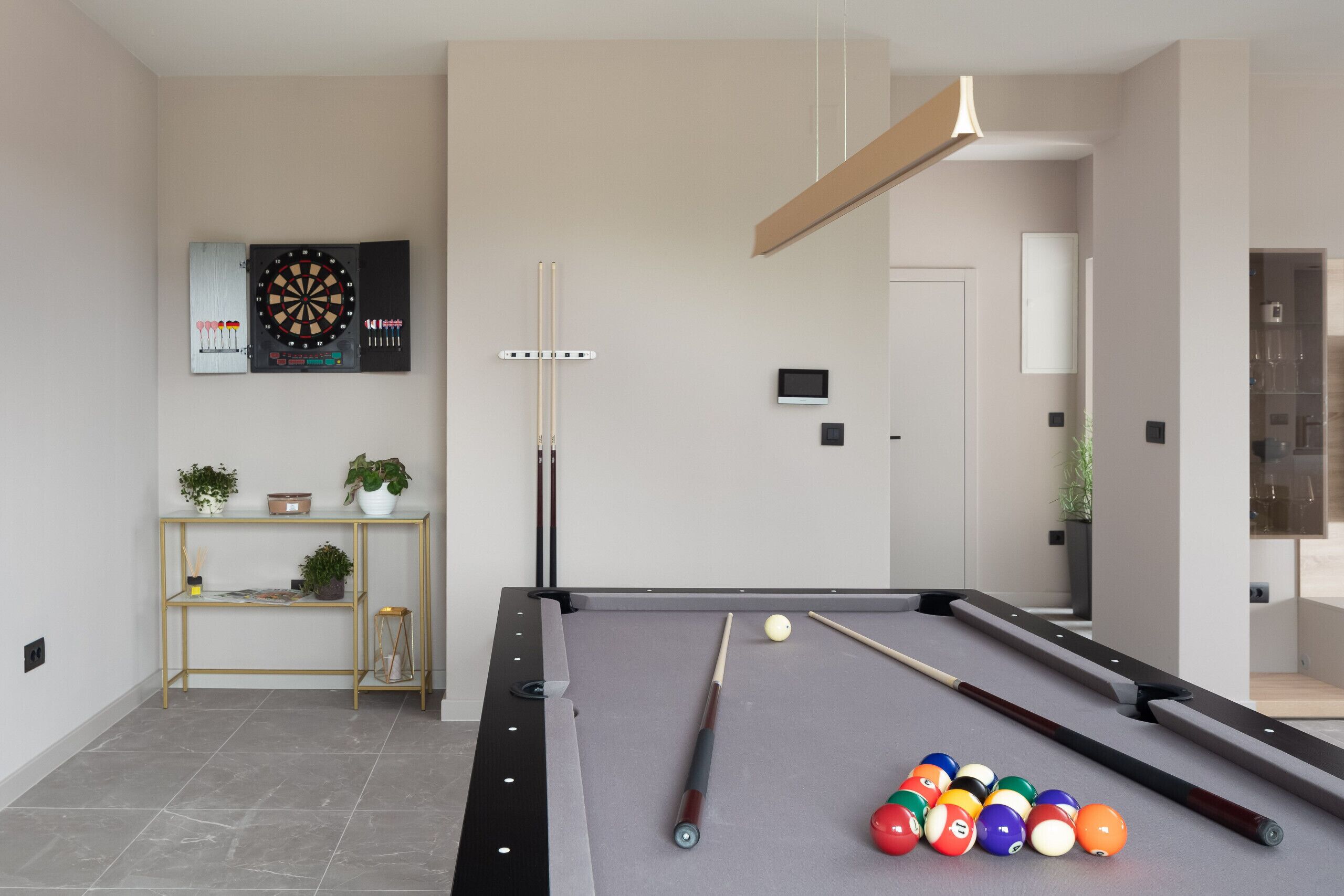 A modern pool table with a dartboard and cue sticks, a small shelf with plants, and a minimalist room design.