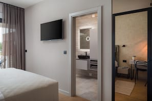 A modern hotel room with a bed, a flat-screen TV mounted on the wall, a bathroom with a sink and mirror, and a small seating area with a chair and table.