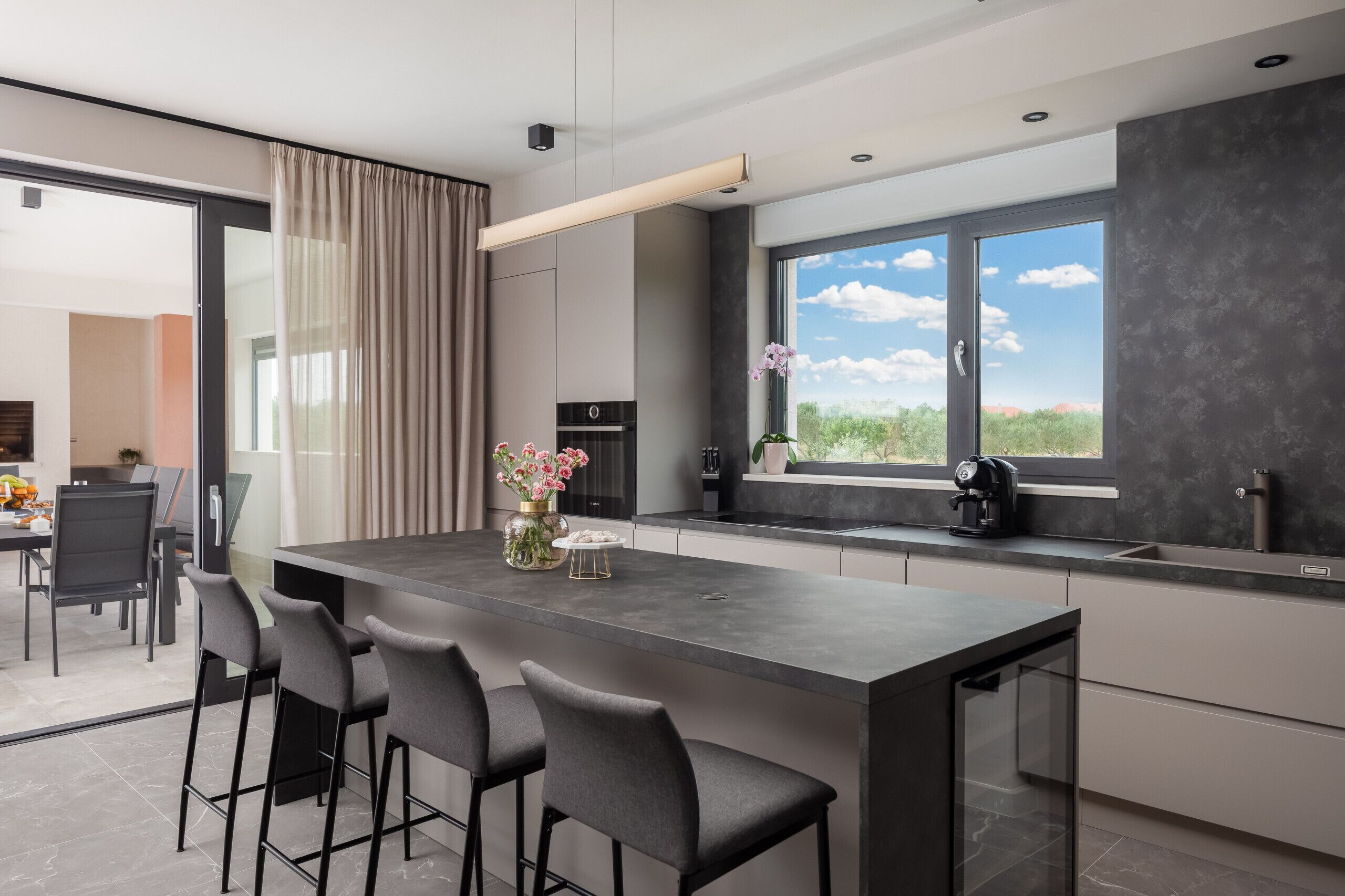 A modern kitchen with a large island, bar stools, a view of the outdoors, and a dining area in the background.