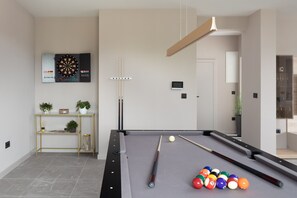 Una mesa de billar moderna con dardos y tacos, un pequeño estante con plantas, y un diseño de habitación minimalista.