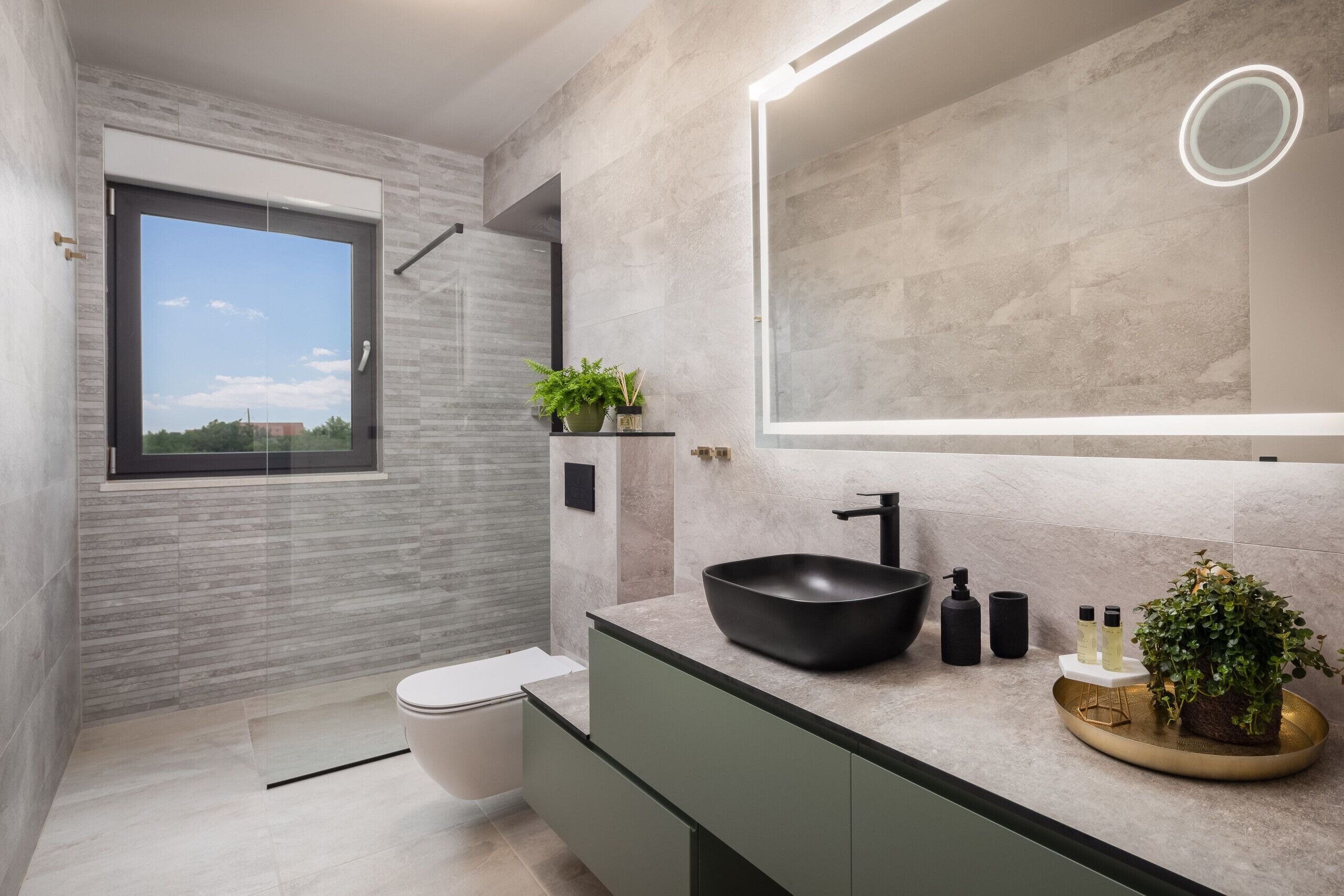 Un bagno moderno con un’ampia finestra, un lavabo nero a incasso, uno specchio con illuminazione integrata e una piccola pianta in vaso sul mobile lavabo.