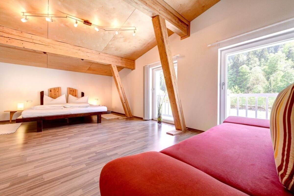 A bedroom with a wooden ceiling, a bed, a red sofa, and a large window with a view of trees.