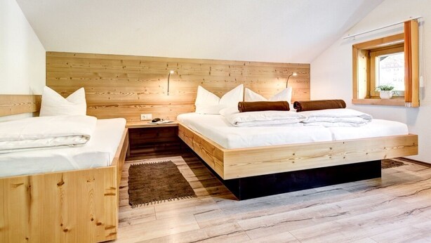 A bedroom with a sloped ceiling, wooden headboard, two beds, a nightstand, a bedside table, a window with a view, and a rug. - Bocks Apartments 61 (Dalaas)