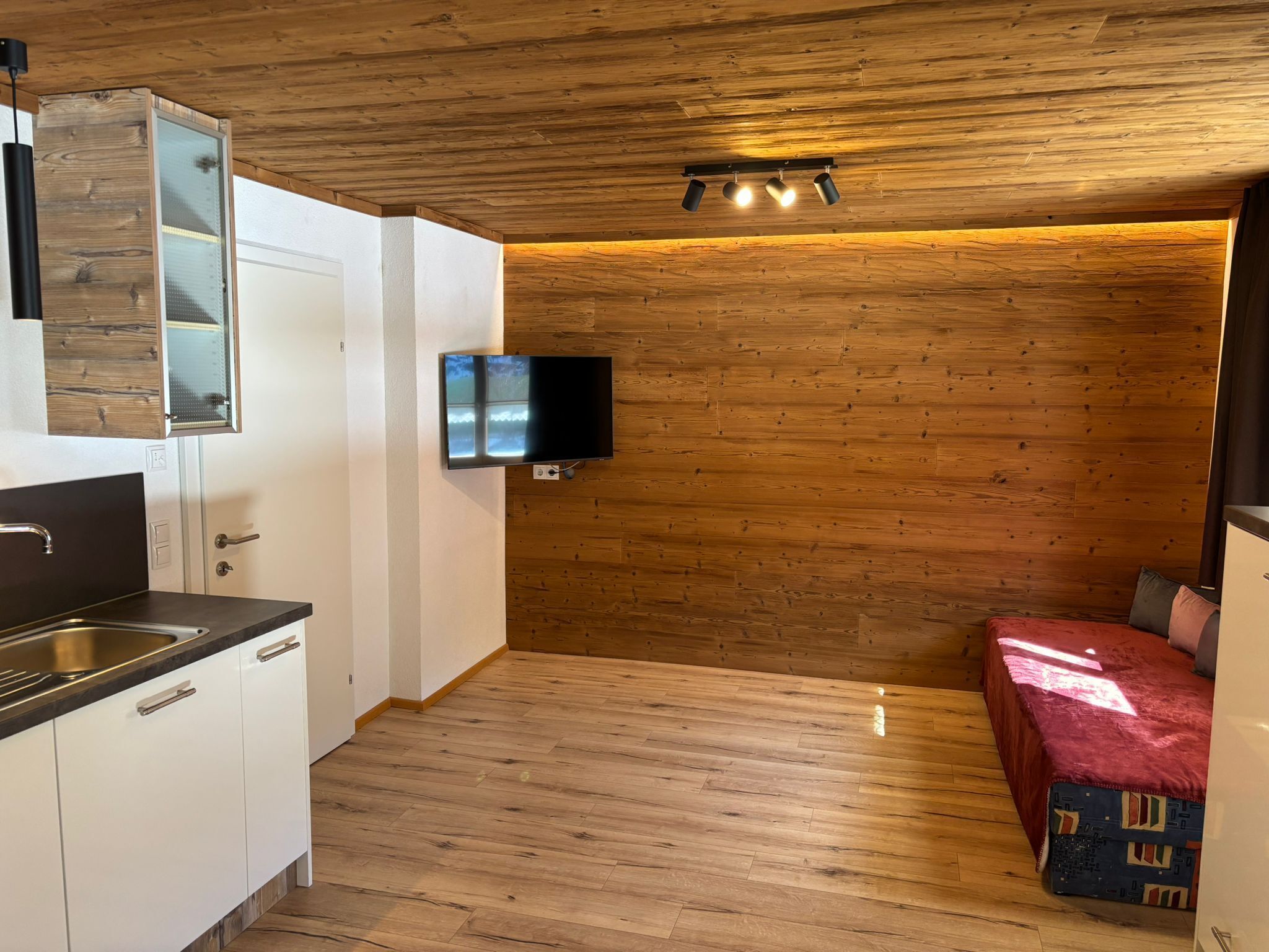 A compact kitchen with a built-in stove, a sink, and a flat-screen TV mounted on the wall.