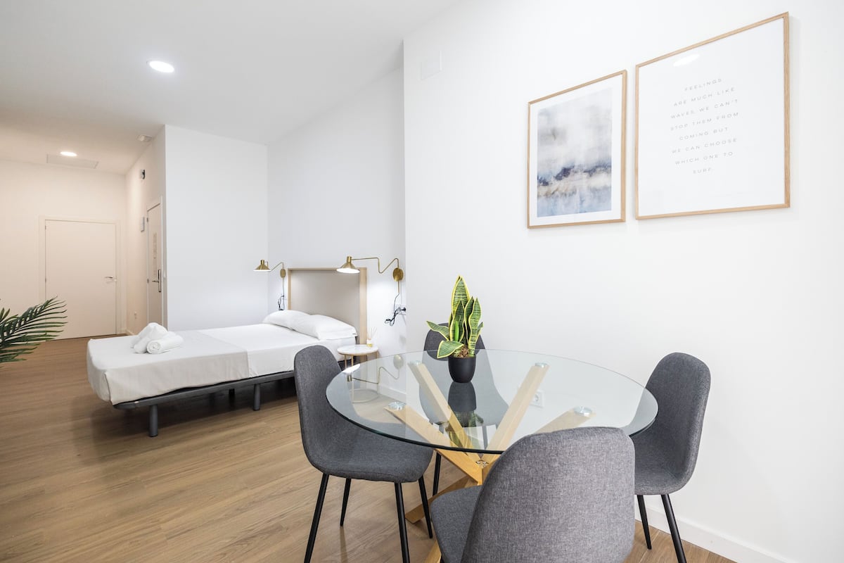 A modern hotel room with a glass dining table, grey upholstered chairs, a bed with white linens, a bedside table with a lamp, and two framed artworks on the wall.