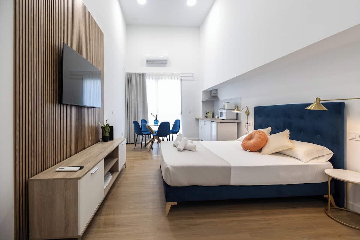 A modern hotel room with a large bed, a TV mounted on a wooden panel wall, a small kitchenette, and a dining area with blue chairs.