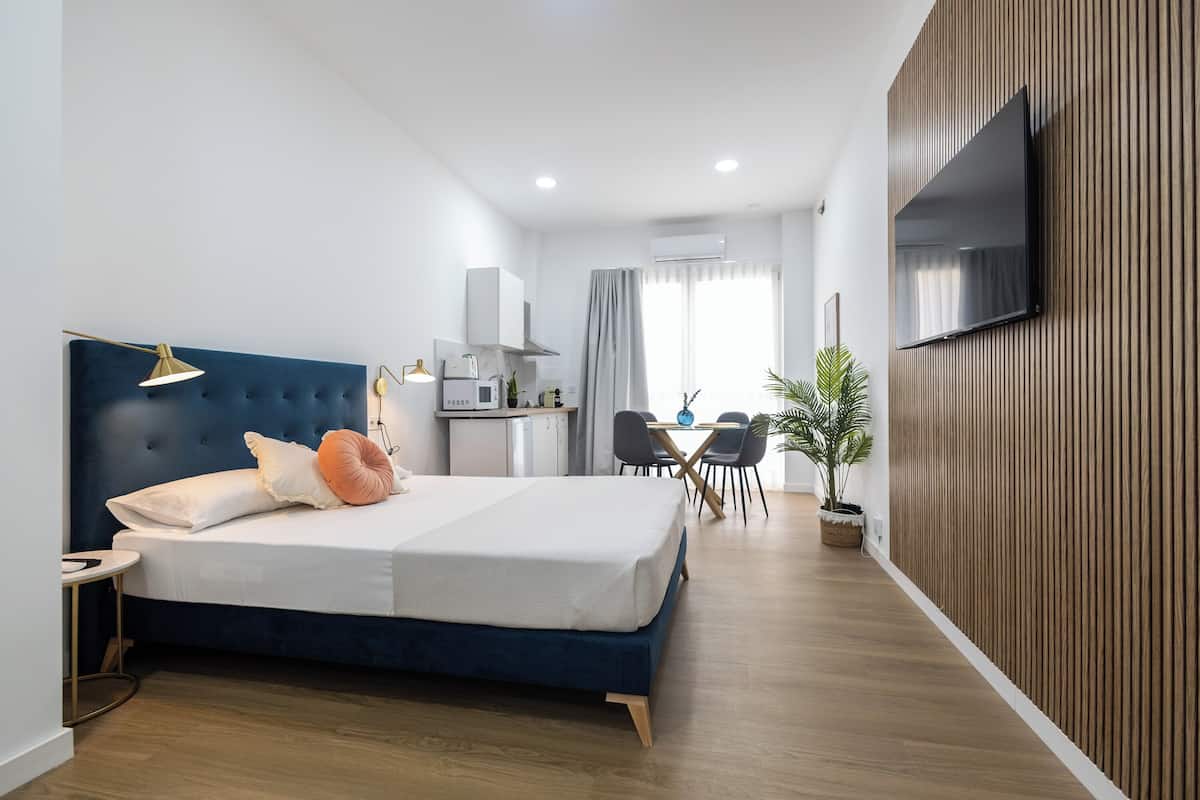 A modern hotel room with a large bed, a desk, a dining area, and a TV mounted on the wall.