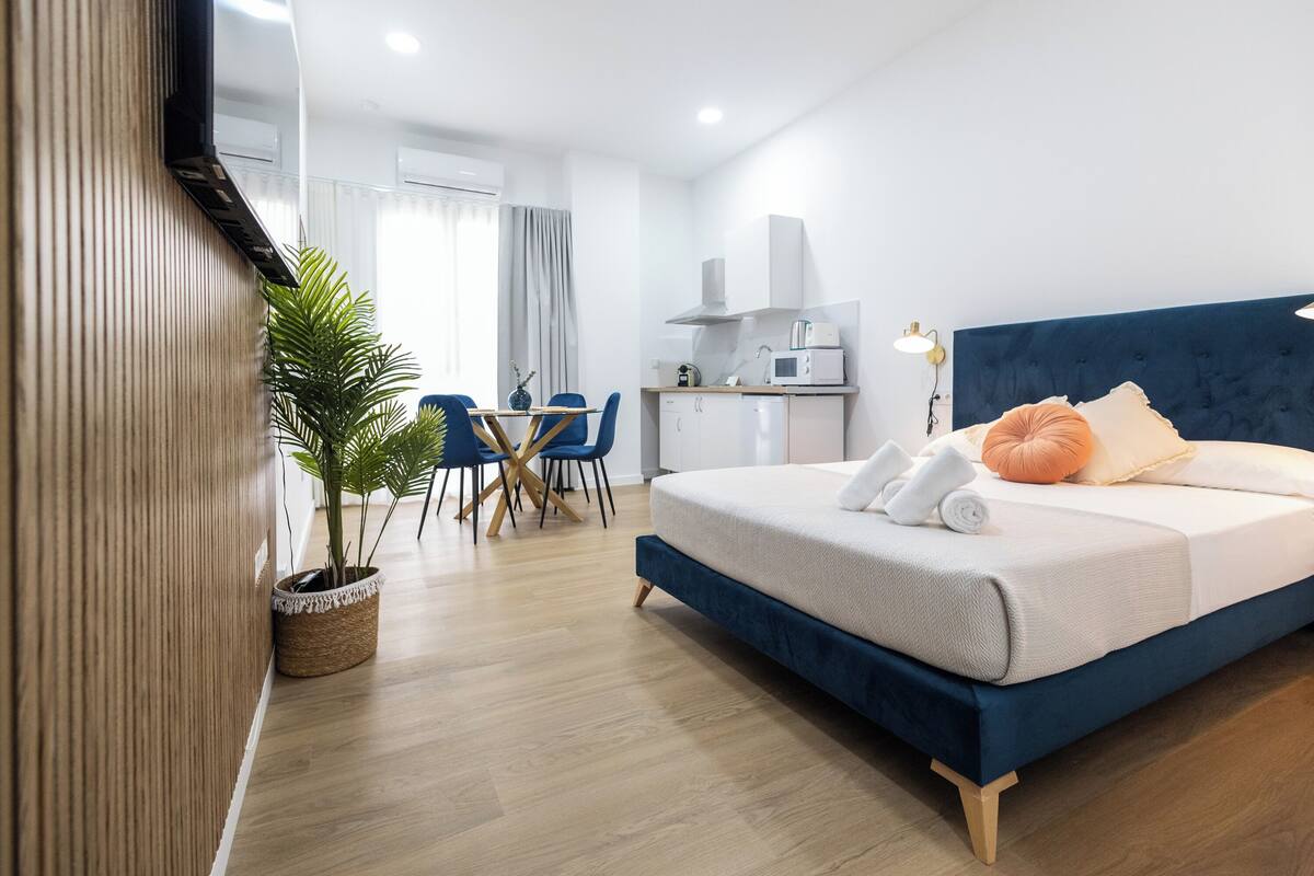A modern hotel room with a bed, a small kitchenette, a dining area, and a potted plant.