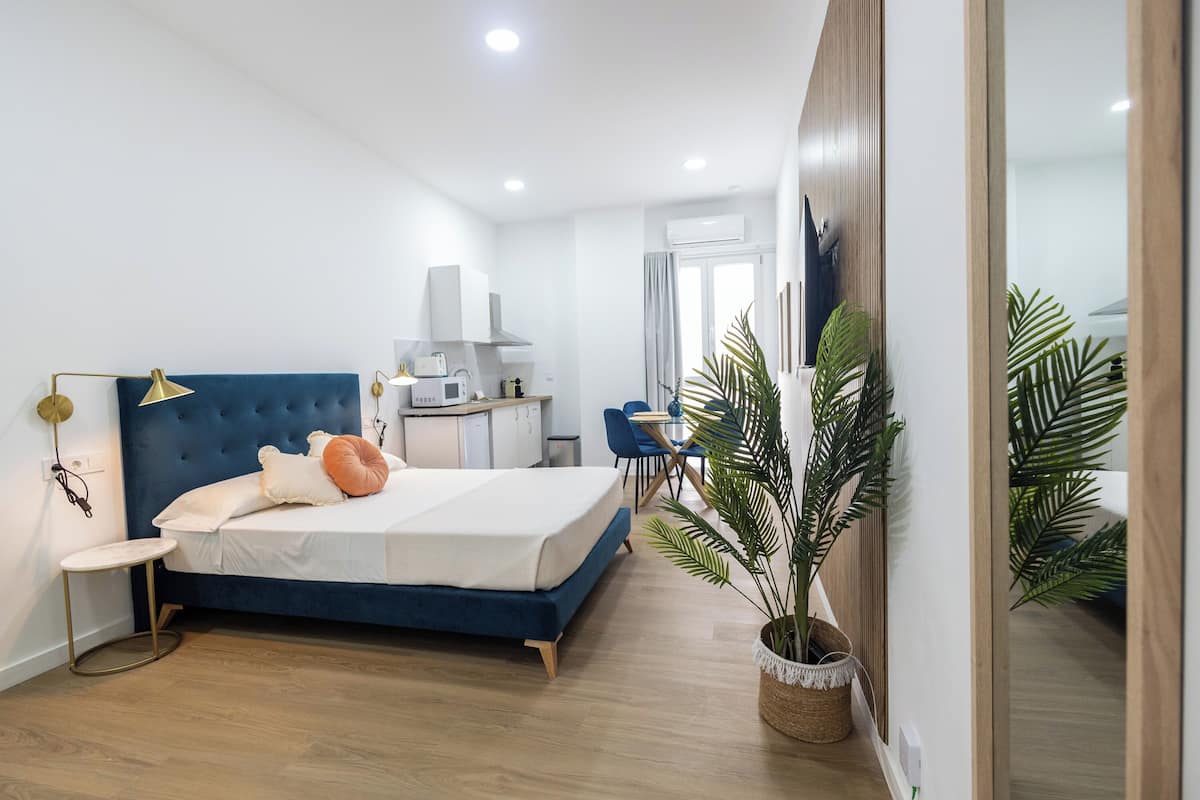 A modern bedroom with a blue headboard, a bedside table with a lamp, a large potted plant, and a view of the kitchen and bathroom through an open door.