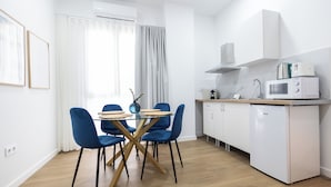 A modern kitchen with white cabinets, a microwave, and a small refrigerator. There is a dining area with a glass table and blue chairs. - Imprenta Sorolla by Concept (València)