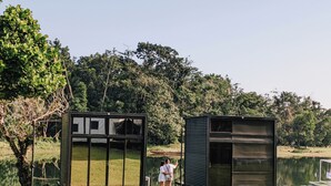 Dos pequeñas estructuras modernas de metal negro con paneles de vidrio, probablemente utilizadas como refugios temporales o unidades de almacenamiento, ubicadas en un área verde con un cuerpo de agua y árboles densos al fondo.
