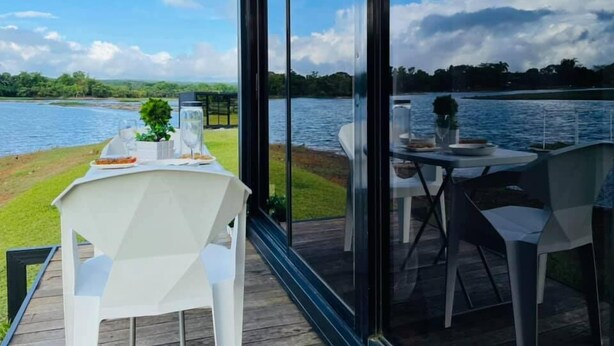 Un comedor con una mesa preparada para dos, con vista a un lago con muelle de césped y barandilla de vidrio.