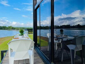 A dining area with a table set for two, overlooking a lake with a grassy deck and a glass railing. - BLOC Camp Site (Cavinti)