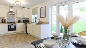 A modern kitchen with white cabinets, a stove, and a microwave. There is a dining area with a table set for four, a vase with pampas grass, and a window with a view of a green lawn. - Modern 4BR House in the Centre Spacious & Ideal for Families and Contractors (Overdale)
