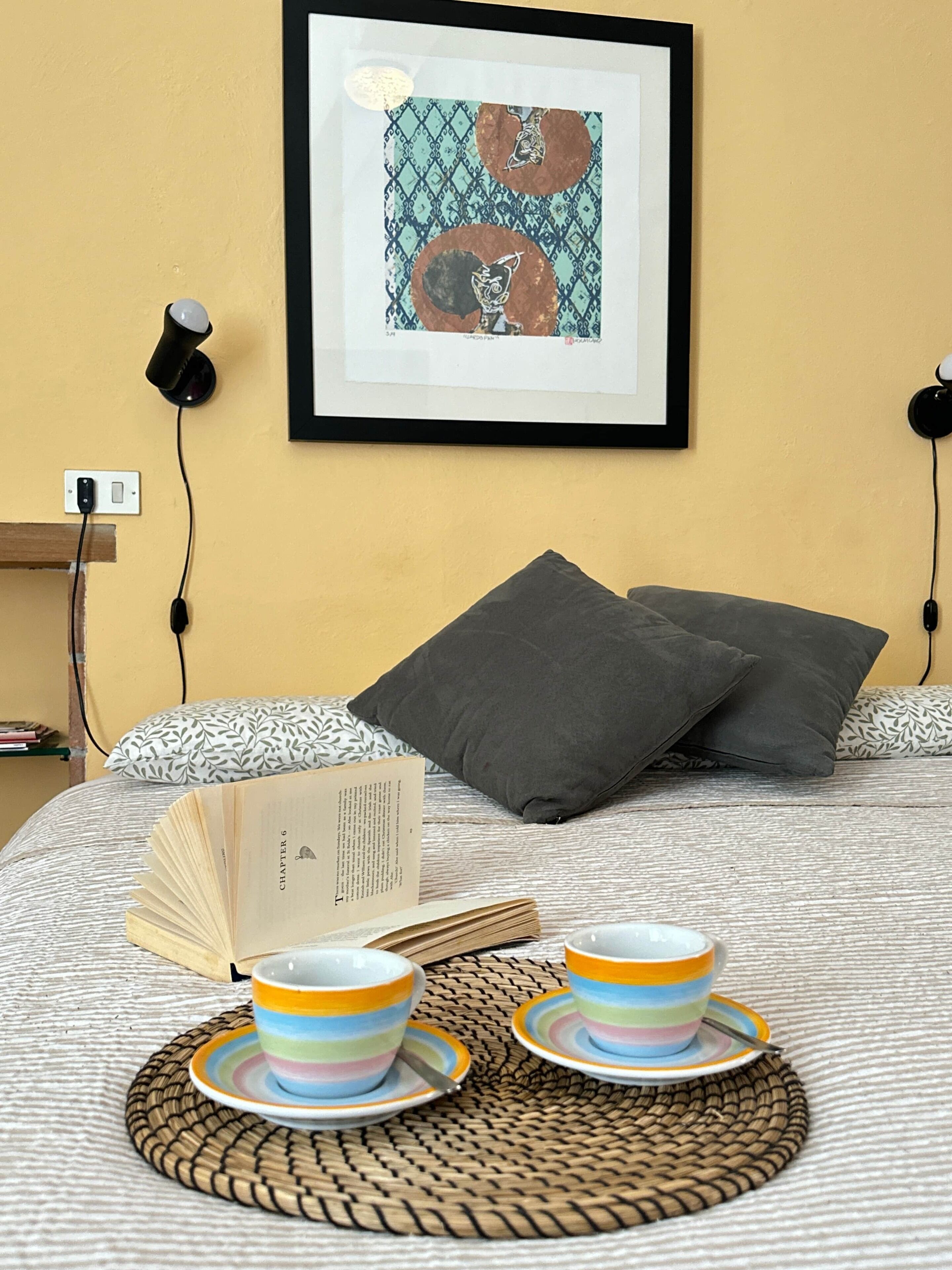 A neatly made bed with a book, two cups, and a framed picture on the wall.