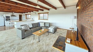 A modern living room with a grey sectional sofa, a wooden coffee table, and a small rug. There is a kitchen area with white cabinets and a microwave. The room features exposed wooden beams and a brick wall.