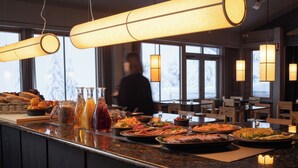 Un buffet proposant divers plats et boissons, une personne à l’arrière-plan et une vue sur l’extérieur à travers de grandes fenêtres.