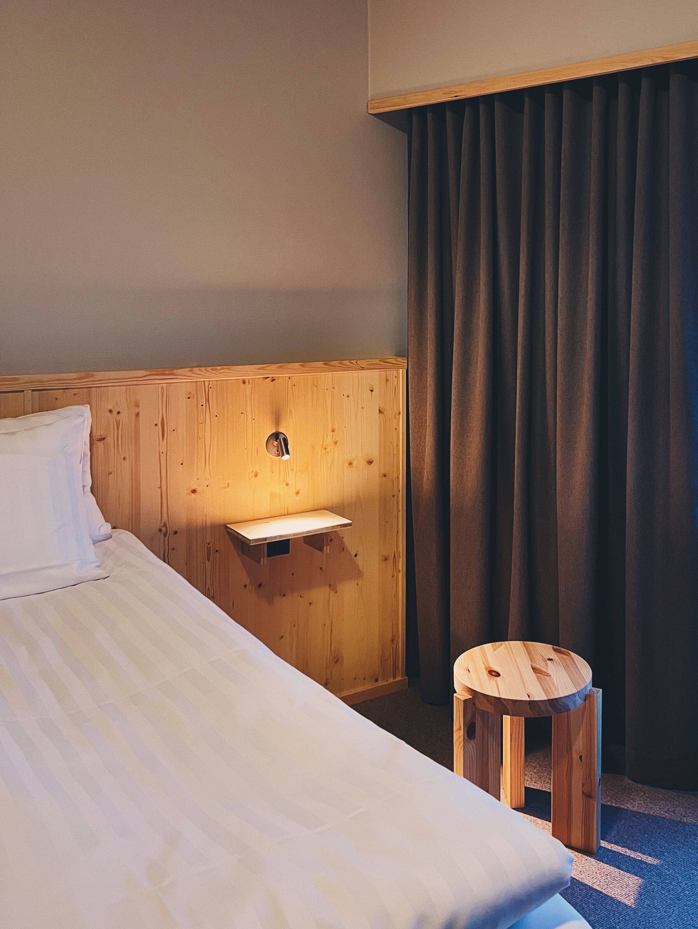 Habitación de hotel con cabecera de madera, un taburete pequeño de madera, una mesita de noche con lámpara y un ventanal con cortinas.