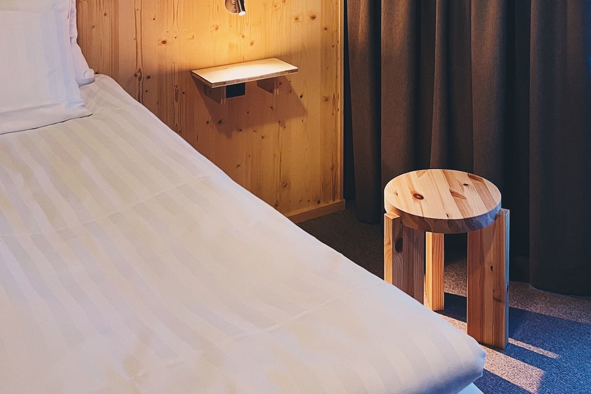 Une chambre d’hôtel avec une tête de lit en bois, un petit tabouret en bois, une table de chevet avec une lampe et une grande fenêtre avec des rideaux.