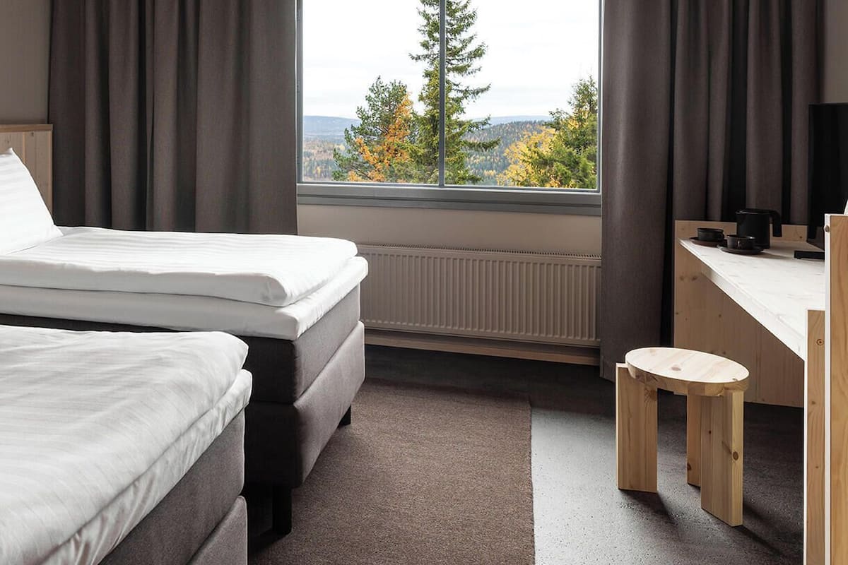 Une chambre d’hôtel avec deux lits, une tête de lit en bois, un bureau avec une chaise et une vue sur les arbres et les collines par la fenêtre.