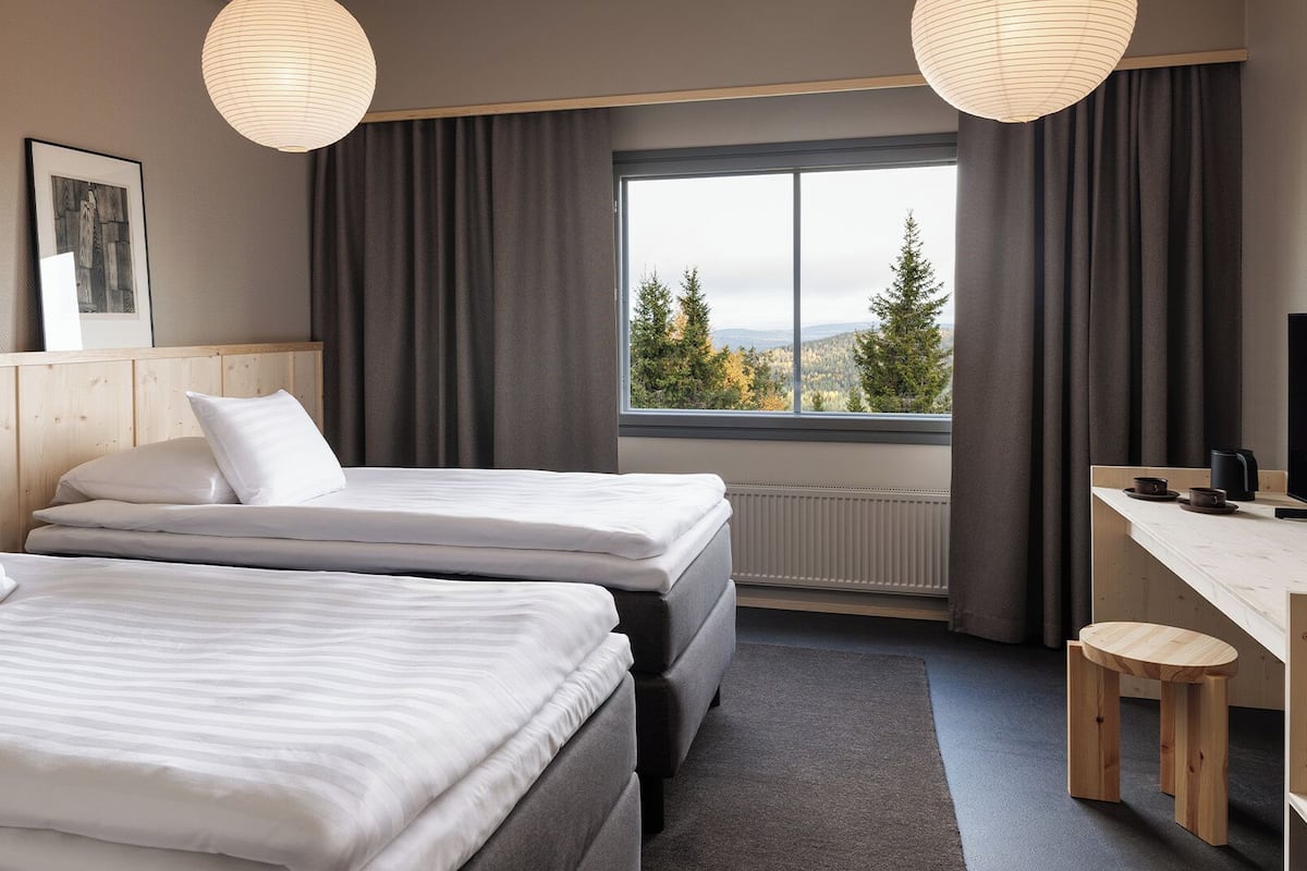 Une chambre d’hôtel avec deux lits, un bureau, une chaise, une fenêtre donnant sur des arbres et des montagnes, et un tableau accroché au mur.
