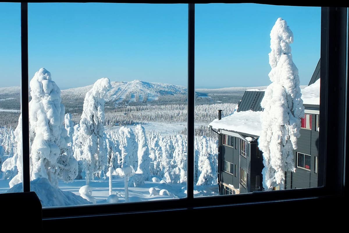 Une vue panoramique d’un paysage enneigé, avec une montagne en arrière-plan, un bâtiment dont le toit est recouvert de neige et des arbres également couverts de neige.