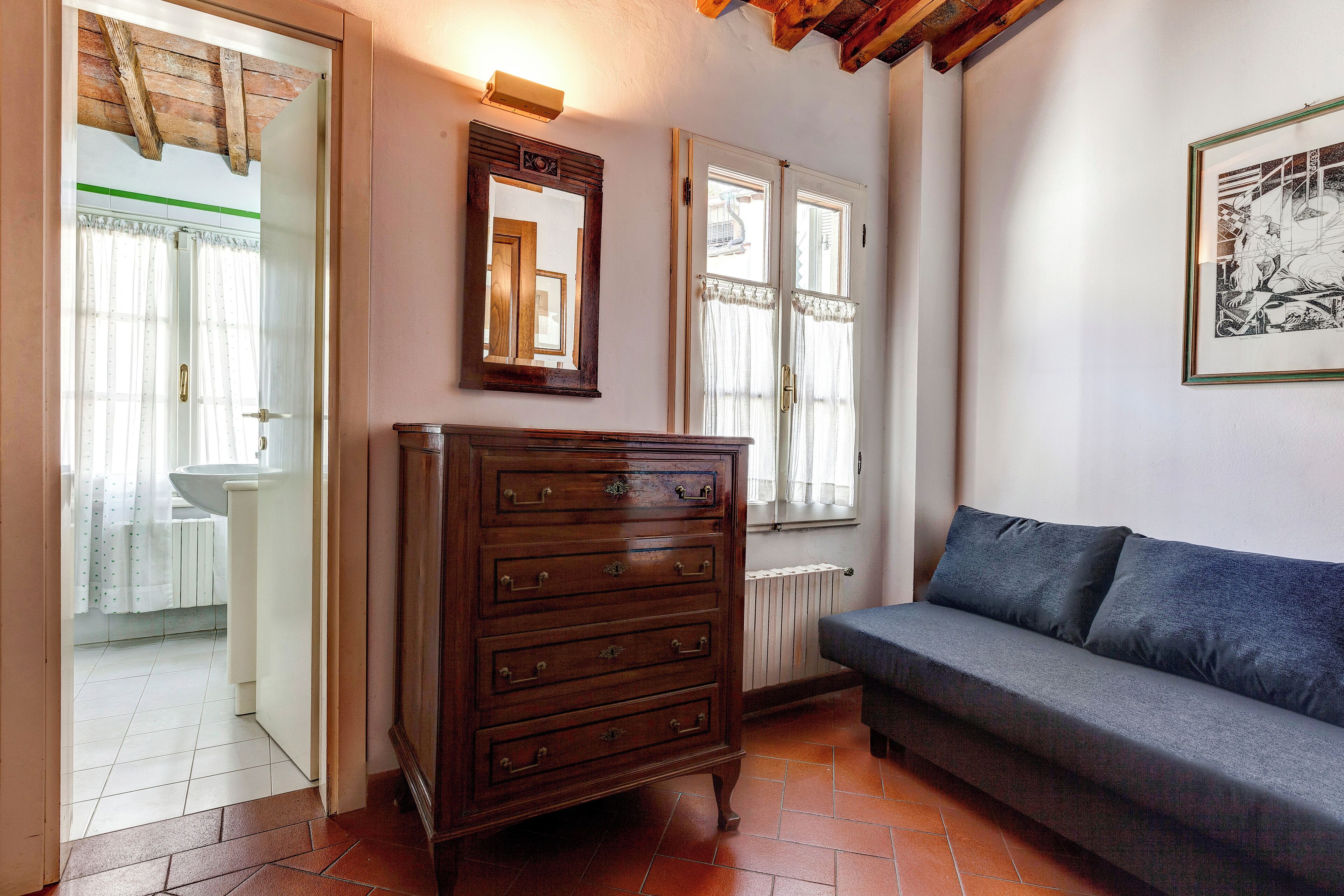 Ein Zimmer mit einem Holzkasten, einem grauen Sofa, einem gerahmten Bild an der Wand und einem Fenster mit Vorhängen.