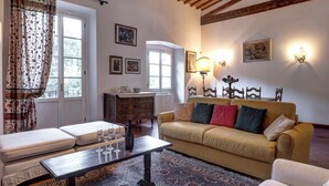 A cozy living room with a sofa, a coffee table, and a patterned rug. There are two windows with curtains, a radiator, and wall-mounted lights. - Farinata Apartment by Mamo Florence (Florence)