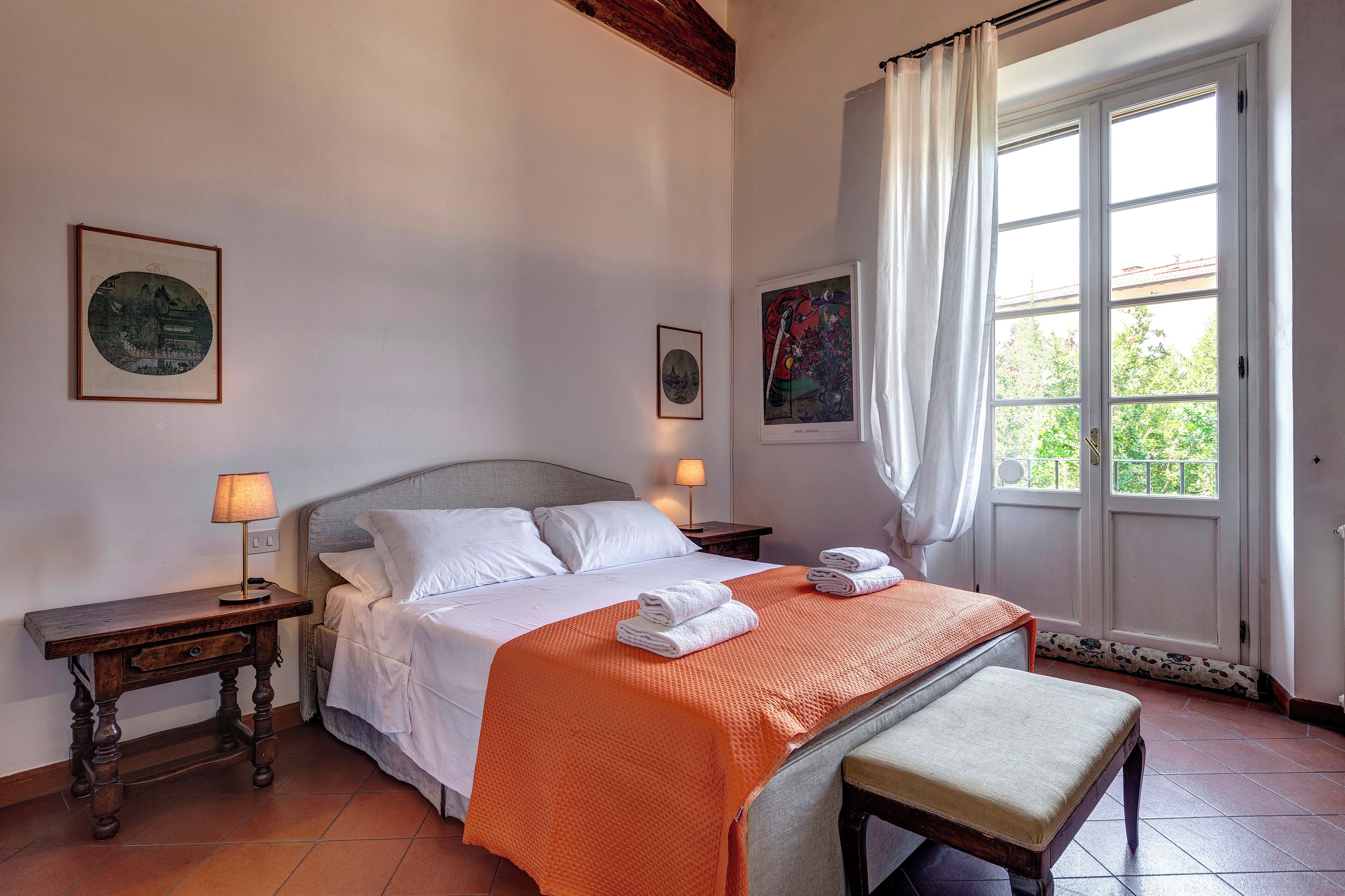Un lit bien fait, avec du linge de lit blanc et une couverture orange, une table de chevet avec une lampe, et une fenêtre avec des rideaux.