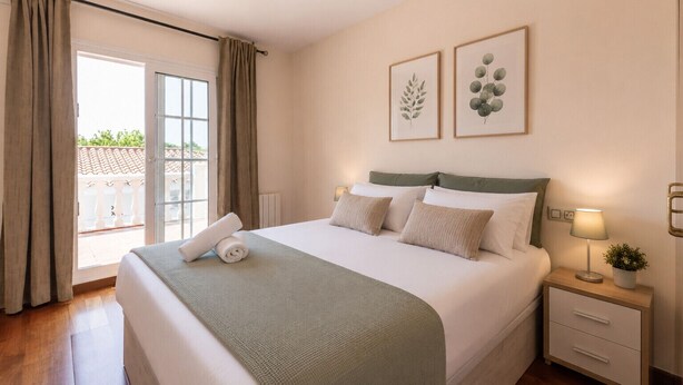 A hotel room with a bed, a nightstand, a lamp, and a view of the outside.