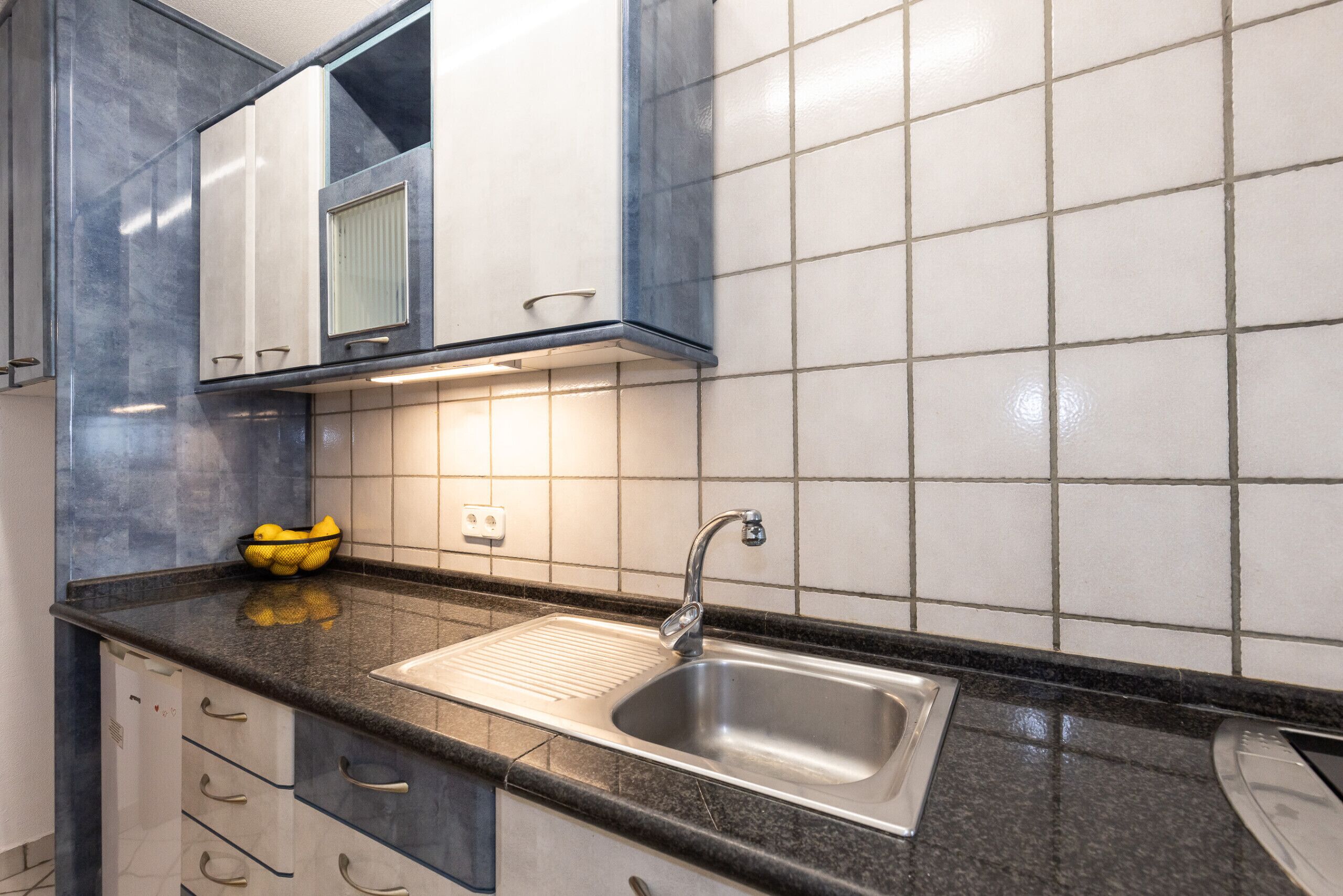 A kitchen with a stainless steel sink, a black countertop, and white tiled walls.