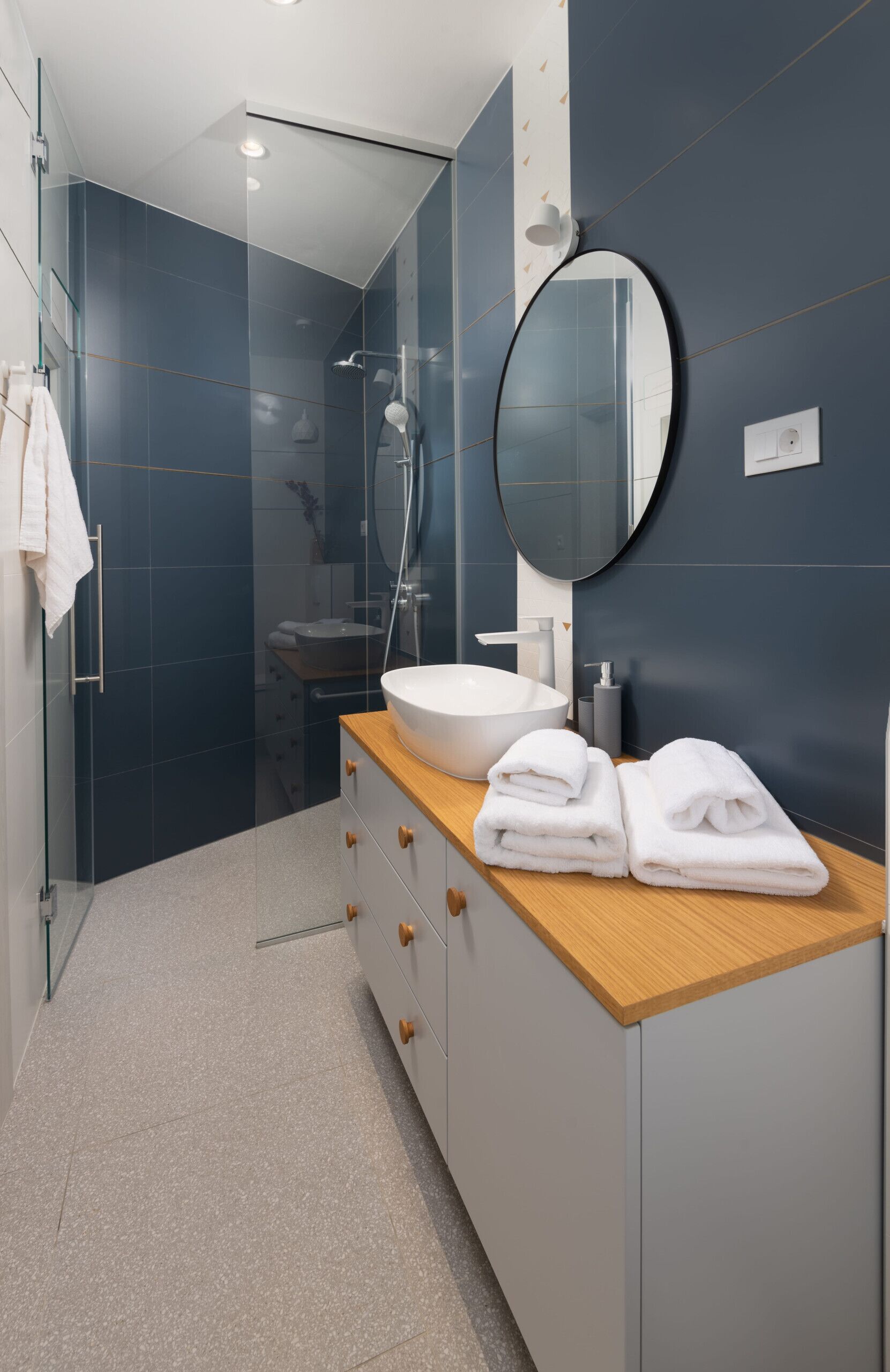A modern bathroom with a glass-enclosed shower, a round mirror, a wooden countertop, and a white sink.