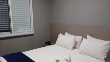 Quarto de hotel com cama, roupa de cama branca, manta azul e janela com persianas.