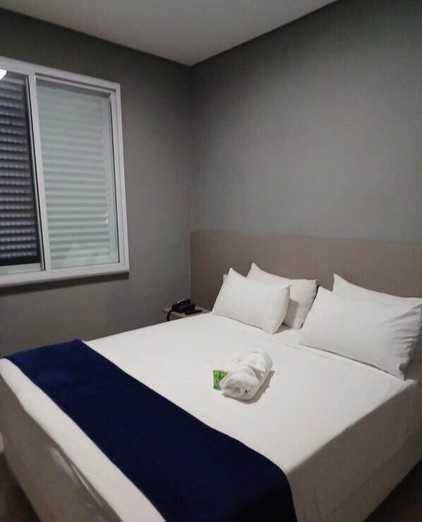 A hotel room with a bed, white bedding, a blue throw blanket, and a window with blinds.