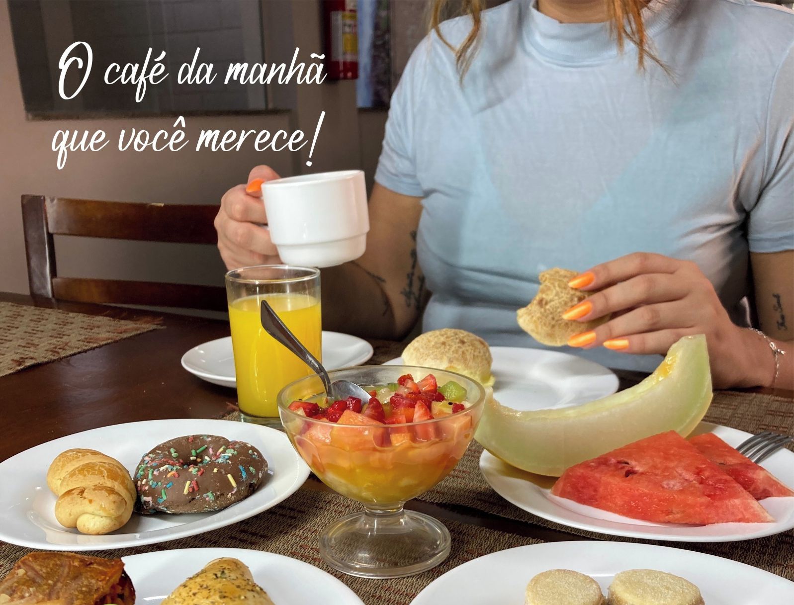 Mesa de café da manhã com diversos doces, um copo de suco de laranja e uma tábua de melancia.