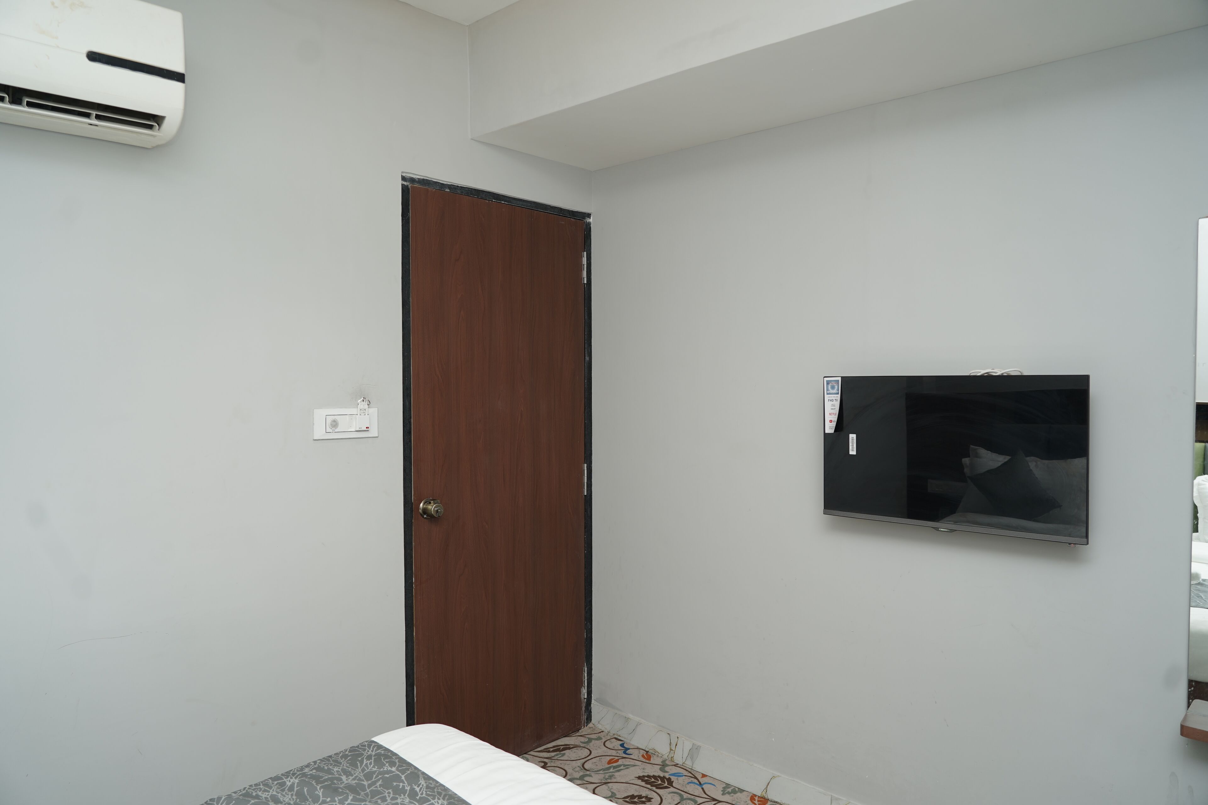 A room with a closed wooden door, a mounted flat-screen TV on the wall, and a ceiling-mounted air conditioner.
